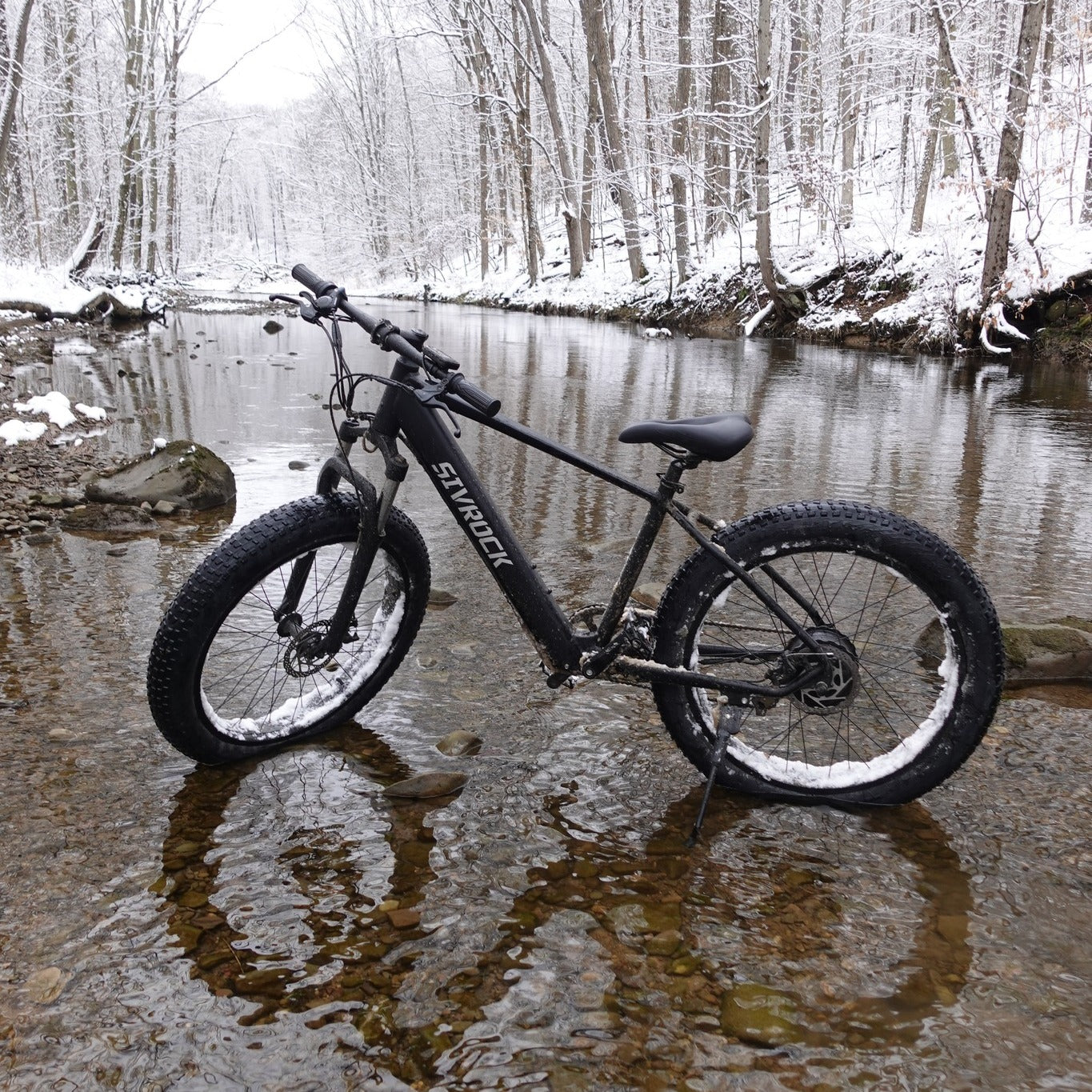 Black electric fat-tire e-bike parked by a river, wide tires and seat visible